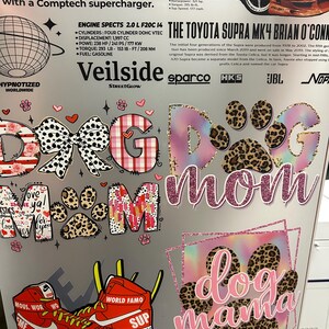 May include: A collage of stickers with the words "Dog Mom" and "Dog Mama" in various fonts and designs. The stickers feature paw prints, bows, hearts, and a pair of red sneakers with the words "World Famo" and "Sup".