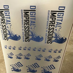 May include: A roll of printed material featuring the text "DIGITAL IMPRESSIONS GRAPHICS & DESIGN" in blue and black. The design includes a stylized lion graphic and the number "10" repeated across the surface. The material is white.