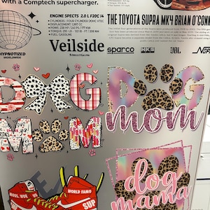 May include: A collage of stickers with car and dog-themed designs. The stickers include the words "Dog Mom" and "Dog Mama" in various fonts and patterns, along with a pair of red sneakers and car specifications.