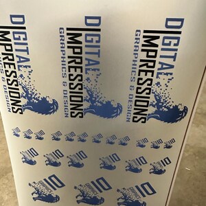 May include: A roll of printed material featuring the text "DIGITAL IMPRESSIONS GRAPHICS & DESIGN" in blue and black. The design includes a stylized lion graphic and the number "10" repeated across the surface. The material is white.