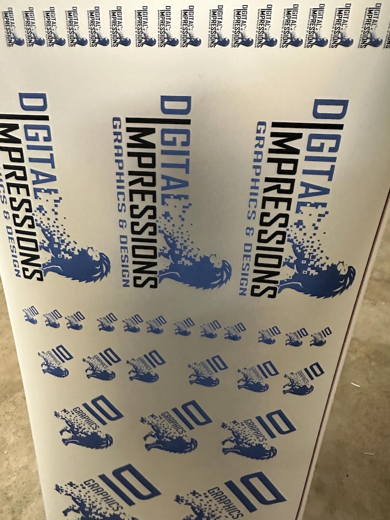 May include: A roll of printed material featuring the text "DIGITAL IMPRESSIONS GRAPHICS & DESIGN" in blue and black. The design includes a stylized lion graphic and the number "10" repeated across the surface. The material is white.