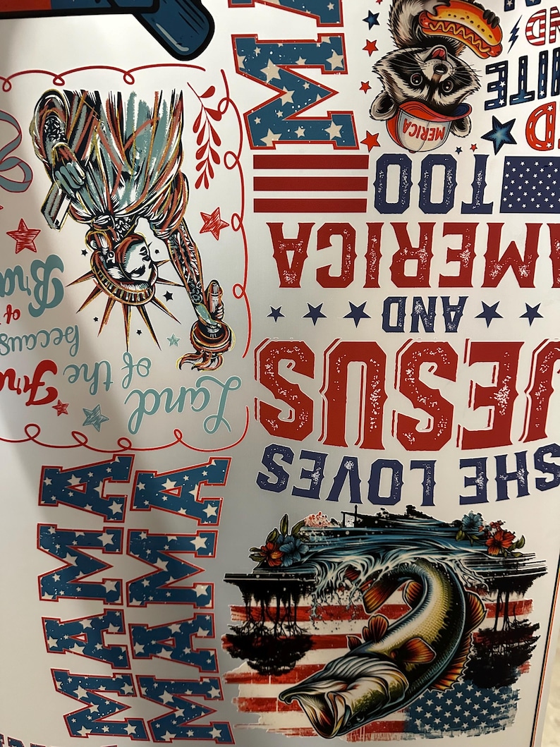 May include: A patriotic design featuring the Statue of Liberty, a raccoon with a hot dog, and a fish leaping from water. The design incorporates the American flag, stars, and text that reads "MAMA", "SHE LOVES JESUS AND AMERICA TOO".