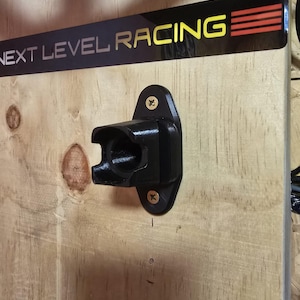 Puede incluir: Soporte de pared de plástico negro con tornillos, fijado a una superficie de madera clara. El soporte de pared tiene un agujero en el centro. Una pegatina negra y amarilla con el texto "NEXT LEVEL RACING" está en la superficie por encima del soporte.
