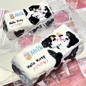 Pode incluir: Duas guloseimas com o tema Hello Kitty, cada uma em uma embalagem plástica transparente. As guloseimas são retangulares com cobertura branca, manchas pretas semelhantes às de vaca e um rosto de Hello Kitty. A palavra "Milk" está escrita em azul, e "Hello Kitty" está escrita em preto com caracteres japoneses.