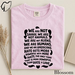 May include: A light pink t-shirt with the text "WE ARE NOT SAVAGE. WE ARE NOT ANIMALS. WE ARE NOT ALIENS. WE ARE HUMANS. AND WE ARE AMERICANS. HATE GETS MORE POWERFUL WITH MORE HATE. THE ONLY THING THAT IS MORE POWERFUL THAN HATE IS LOVE."