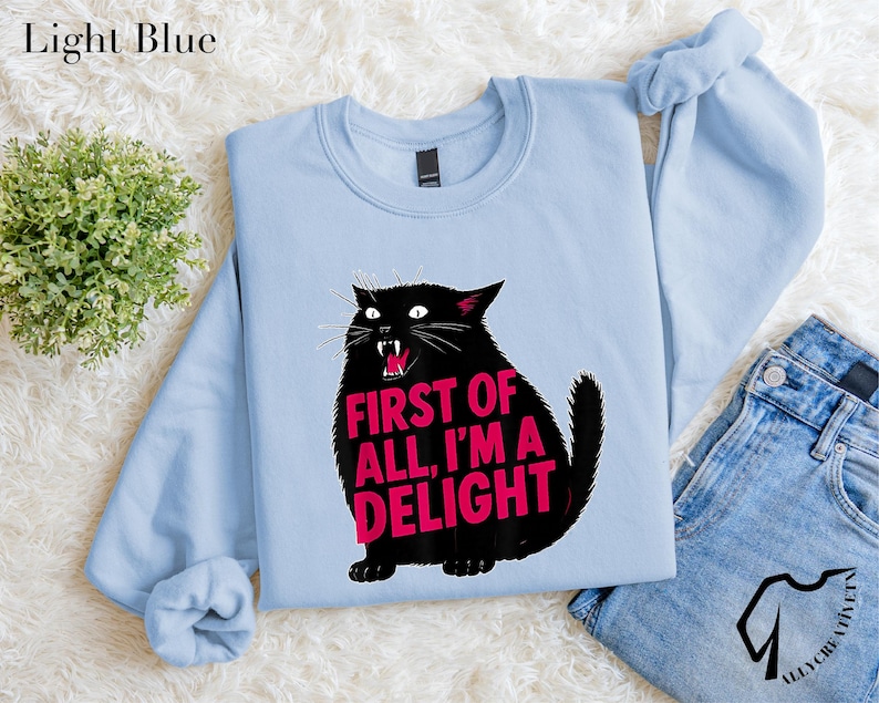 A light blue sweatshirt with a graphic of a black cat and the text "FIRST OF ALL, I'M A DELIGHT" printed on it. The sweatshirt is laid out on a white surface, along with a pair of jeans and a potted plant.