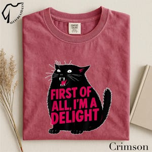 A red t-shirt with a graphic design of a black cat and the text "FIRST OF ALL, I'M A DELIGHT" printed on it.