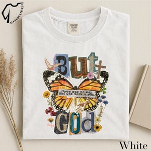 Puede incluir: Camiseta blanca con un diseño gráfico colorido. El diseño presenta la palabra "BUT" en letras azules y marrones, una mariposa, una cruz y las palabras "there was no way, but God made a way." La camiseta también tiene el texto "GOD" en letras coloridas.