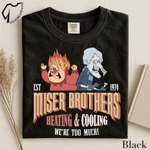 May include: A black crew neck t-shirt featuring a graphic of the Miser Brothers with the text EST 1974 MISER BROTHERS HEATING & COOLING WE'RE TOO MUCH. The design shows Heat Miser in red and Snow Miser in blue, printed on a Comfort Colors brand shirt.