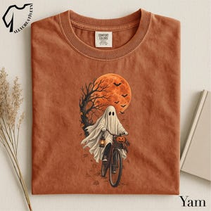 May include: A rust-colored crew neck t-shirt featuring a graphic of a ghost riding a bicycle. The ghost carries a lantern and a pumpkin in the bike basket. A large orange moon, a bare tree, and flying bats are in the background with falling leaves.