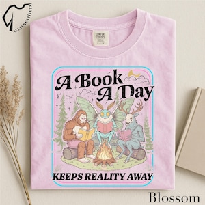 May include: A light pink t-shirt featuring a graphic of a Bigfoot, a moth, and a deer-like creature sitting around a campfire reading books. The text reads A BOOK A DAY KEEPS REALITY AWAY. The design includes pine trees, clouds, and a crescent moon.