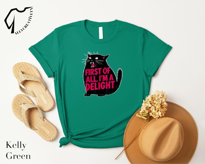 a green t-shirt with a cat graphic and the text "FIRST OF ALL I'M A DELIGHT" printed on it, along with a pair of beige flip-flops and a tan hat.