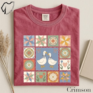 May include: Crimson-colored t-shirt featuring a patchwork design with floral and geometric patterns. The central square showcases two white ducks with blue bows. The shirt has a crew neck and the text "Crimson" at the bottom.