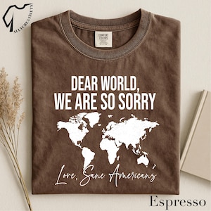 May include: Espresso-colored t-shirt with the text "DEAR WORLD, WE ARE SO SORRY" and a white world map graphic. The shirt also includes the phrase "Love, Sane Americans" in a script font. The shirt is a Comfort Colors brand.
