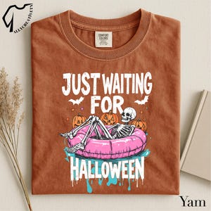 May include: A rust-colored t-shirt featuring a graphic of a skeleton lounging in a pink pool float. The text reads "JUST WAITING FOR HALLOWEEN" in white letters. The design includes orange pumpkins, small bats, and blue water accents below the float.