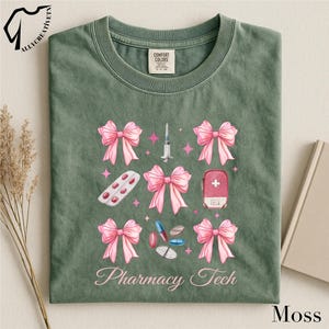 May include: Moss green t-shirt with a crew neck. The shirt features a design of pink bows, pills, a syringe, a first aid kit, and the words "Pharmacy Tech" in a script font. The shirt is made of a soft, comfortable material.