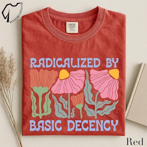 May include: A red t-shirt with the words "RADICALIZED BY BASIC DECENCY" in blue lettering. The shirt features a floral design in pink, orange, and green. The shirt is folded neatly.
