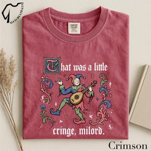 May include: A crimson crew neck t-shirt featuring a graphic of a jester playing a lute, surrounded by ornate floral scrollwork. The text reads, "That was a little cringe, milord." The shirt has a Comfort Colors label at the inner neckline.