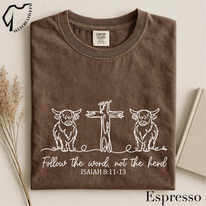 May include: Espresso-colored t-shirt with a white graphic of a cross and two Highland cows. The text "Follow the word, not the herd" and Bible verse Isaiah 8:11-13 are printed below the design. The shirt is a Comfort Colors brand.