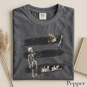 May include: A charcoal gray t-shirt featuring a graphic of a skeleton holding a shotgun and a mallard duck flying away with a skeletal arm. The text "Well, shit..." is printed below the duck. The shirt has a Comfort Colors brand label at the neck.