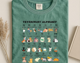 May include: A sage green t-shirt featuring a black graphic print titled Veterinary Alphabet. The design displays letters A through Z, each paired with a small illustration of animals or veterinary themes and corresponding text labels.