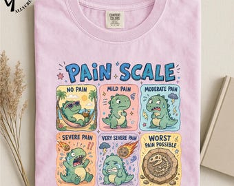 May include: A light pink t-shirt featuring a graphic titled PAIN SCALE with six panels showing a green dinosaur in various states. The panels depict no pain, mild pain, moderate pain, severe pain, very severe pain, and worst pain possible.