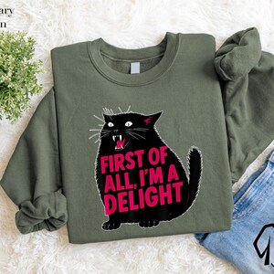 a green sweatshirt with a graphic design of a black cat and the text "FIRST OF ALL, I'M A DELIGHT" printed on it. The sweatshirt is placed on a white surface, with a pair of blue jeans visible in the background.