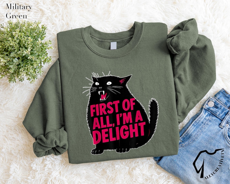 a green sweatshirt with a graphic design of a black cat and the text "FIRST OF ALL, I'M A DELIGHT" printed on it. The sweatshirt is placed on a white surface, with a pair of blue jeans visible in the background.