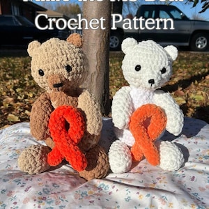 May include: Two crocheted teddy bears, one tan and one white, each holding an orange ribbon. The bears are sitting on a floral patterned blanket. Text at the top reads "Millie the MS Bear Crochet Pattern."