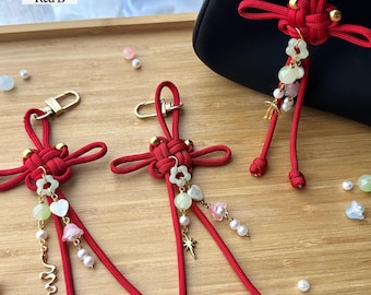 May include: Three red cord keychains featuring intricate knot designs, gold-tone lobster clasps, and dangling charms including small flower-shaped beads, heart-shaped beads, pearls, and metallic accents like a star and a snake. Text reads Red B.