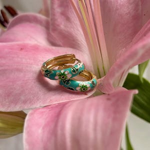 Könnte beinhalten: Ein Paar goldfarbene Creolen mit einem türkisfarbenen Emaille-Blumenmuster, die auf einem rosa Lilienblatt liegen. Die Ohrringe weisen ein detailliertes Muster aus weißen und grünen Blumen auf. Der Hintergrund ist unscharf.