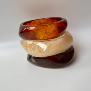 Könnte beinhalten: Drei übereinander gestapelte Armreifen aus Harz. Der obere Armreif ist bernsteinfarben, der mittlere cremefarben und der untere dunkelbraun. Die Armreifen haben eine glatte, abgerundete Form und sind aus Harz gefertigt.