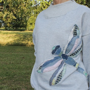 May include: A light gray sweatshirt featuring a large, decorative dragonfly appliqué on the front. The dragonfly is constructed from various fabric scraps, including blue denim, plaid, and striped patterns with frayed, raw edges.