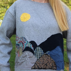 Puede incluir: Una sudadera gris jaspeado de cuello redondo con un parche circular amarillo que recuerda a un sol y una escena de paisaje en la parte inferior frontal, construida con varios parches de tela en azul marino, azul, cuadros y flores con costuras.
