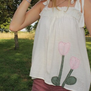 May include: A white sleeveless top featuring a gathered neckline with tie-up shoulder straps. The garment is decorated with two embroidered pink tulip flowers with green stems and leaves positioned on the lower right side of the front panel.