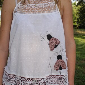 May include: A white sleeveless top featuring a square neckline with intricate white lace trim, a dotted fabric texture, and two appliquéd ladybug figures with black heads and red-and-black patterned wings, finished with a wide scalloped lace hem.