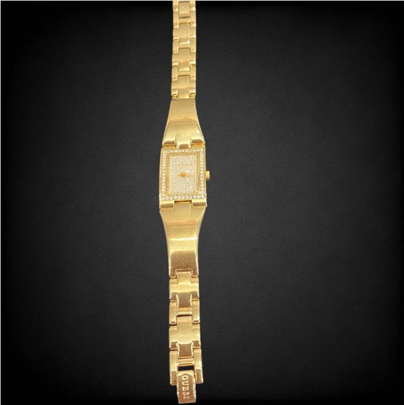 Pu&ograve; includere: Un orologio Guess dorato con un quadrante quadrato e un design a cristalli simili a diamanti. L'orologio ha un cinturino in metallo dorato.