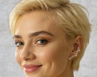 Può includere: Una donna con i capelli biondi corti sorride, mostrando un ear cuff decorativo. L'ear cuff presenta un design floreale con piccoli abbellimenti scintillanti. Lo sfondo è un grigio morbido e neutro.