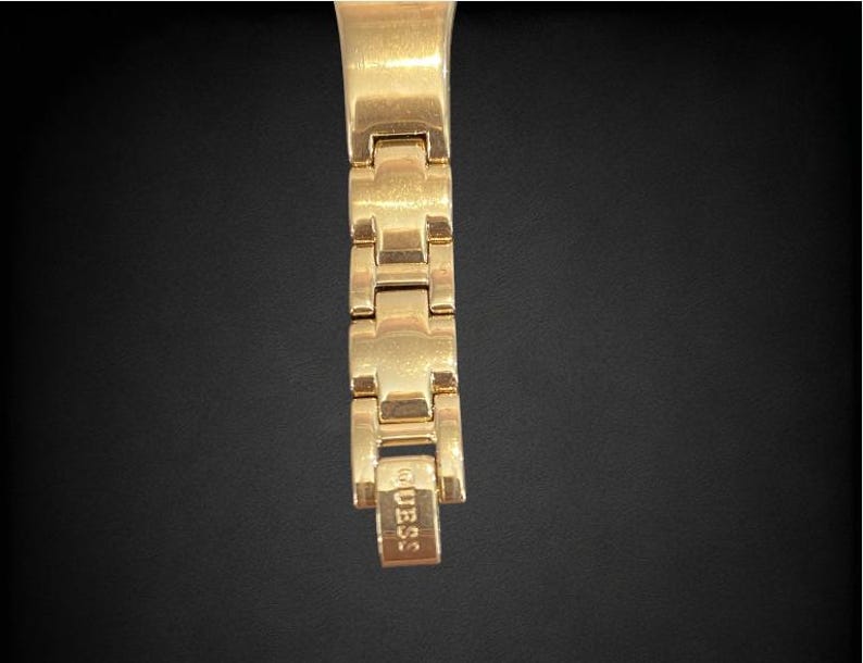 Pu&ograve; includere: Cinturino da orologio dorato con una chiusura che recita "GUESS".
