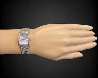 Può includere: Un orologio argentato con quadrante quadrato e cinturino in maglia argentata. Il quadrante dell'orologio ha uno sfondo viola chiaro con lancette e indici argentati.