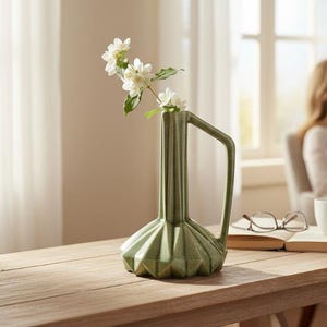 May include: A sage green ceramic vase with a faceted, geometric base and a tall, slender neck featuring a single angular handle. The vase has a glossy, textured finish and contains a delicate sprig of white jasmine flowers with green leaves.
