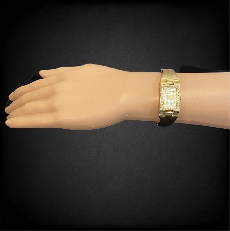 Pu&ograve; includere: Un orologio color oro con un quadrante rettangolare e una lunetta tempestata di diamanti. L'orologio &egrave; su un polso.