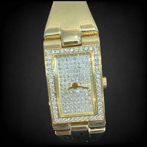 Pu&ograve; includere: Un orologio da polso Guess color oro con un quadrante rettangolare e una lunetta tempestata di diamanti. Il quadrante dell'orologio &egrave; anche ricoperto di piccoli diamanti e presenta la parola "Guess" al centro.
