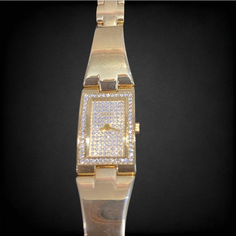 Pu&ograve; includere: Un orologio Guess dorato con un quadrante rettangolare e una lunetta tempestata di cristalli. L'orologio ha un cinturino in metallo dorato.