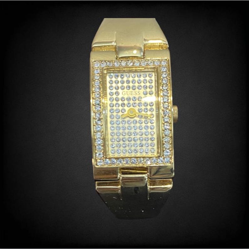 Pu&ograve; includere: Un orologio Guess color oro con un quadrante rettangolare e un cinturino composto da pezzi di metallo collegati. Il quadrante &egrave; ricoperto da piccoli strass trasparenti.