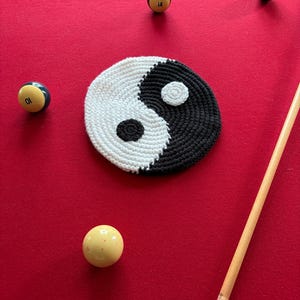 Puede incluir: Un posavasos redondo hecho a mano de ganchillo con un diseño de yin-yang, con secciones en blanco y negro. El posavasos está sobre una superficie roja, rodeado de bolas de billar y un taco. El posavasos tiene aproximadamente 10 cm de diámetro.