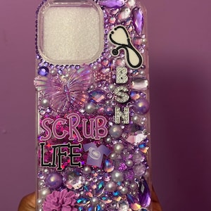May include: A clear phone case bedazzled with purple and silver rhinestones, pearls, and decorative elements. The case features the text "SCRUB LIFE" and a stethoscope charm, with the letters "BSN" in silver. A purple flower and butterfly add to the design.