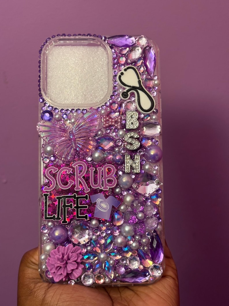 May include: A clear phone case bedazzled with purple and silver rhinestones, pearls, and decorative elements. The case features the text "SCRUB LIFE" and a stethoscope charm, with the letters "BSN" in silver. A purple flower and butterfly add to the design.