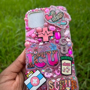 May include: A pink bedazzled phone case with the letters "ICU" in leopard print. The case features medical-themed charms, including a first aid kit, pills, and a heartbeat symbol. The words "Scrub Life" are also visible.