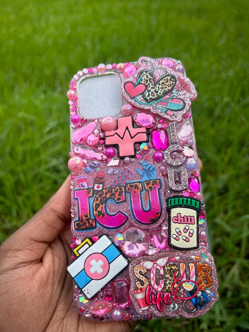 May include: A pink bedazzled phone case with the letters "ICU" in leopard print. The case features medical-themed charms, including a first aid kit, pills, and a heartbeat symbol. The words "Scrub Life" are also visible.